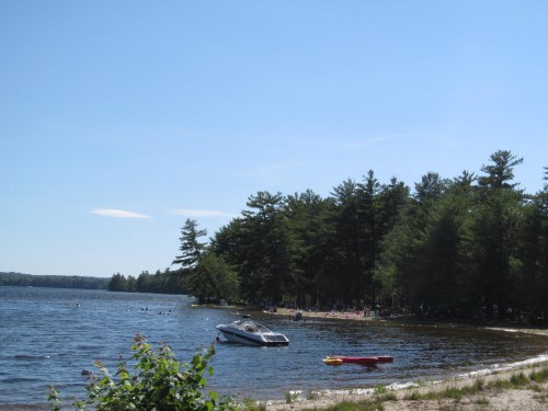 A serene lake scene with a boat on the water, surrounded by trees and a sandy beach with people enjoying the sun.