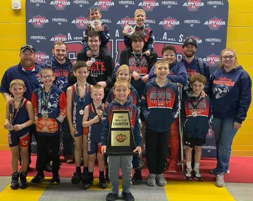 A group of young wrestlers and coaches pose together, holding a championship plaque and wearing medals, celebrating their win.