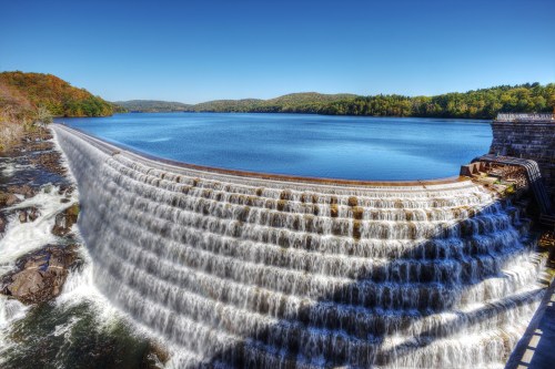 A scenic view of a dam with cascading water, surrounded by lush greenery and a clear blue sky.
