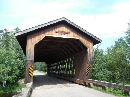 A wooden covered bridge with a peaked roof, surrounded by trees and a clear blue sky.