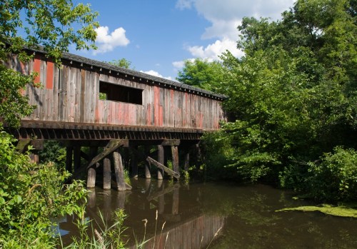 A wooden covered bridge stands over a calm, green-tinted pond, surrounded by lush trees and blue skies.