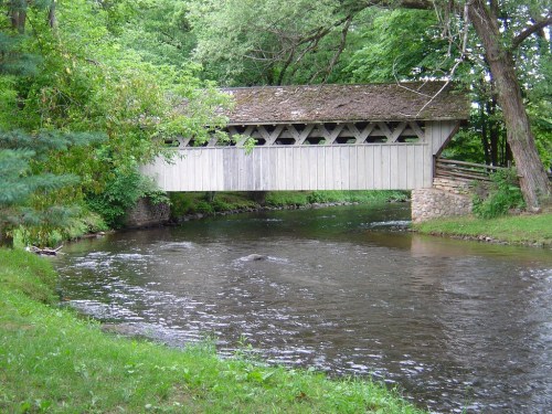 A wooden covered bridge spans a calm river, surrounded by lush green trees and grass.