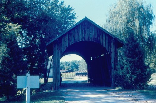 A wooden covered bridge surrounded by trees, leading to a path and open area beyond.