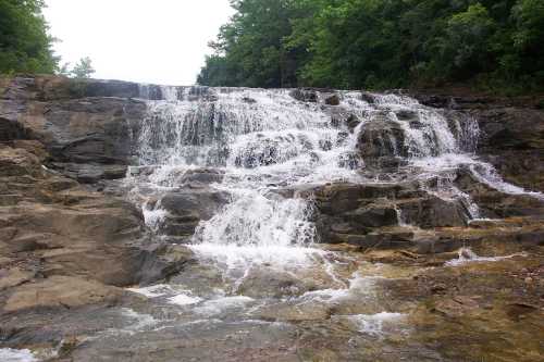 A cascading waterfall flows over rocky terrain, surrounded by lush green trees.