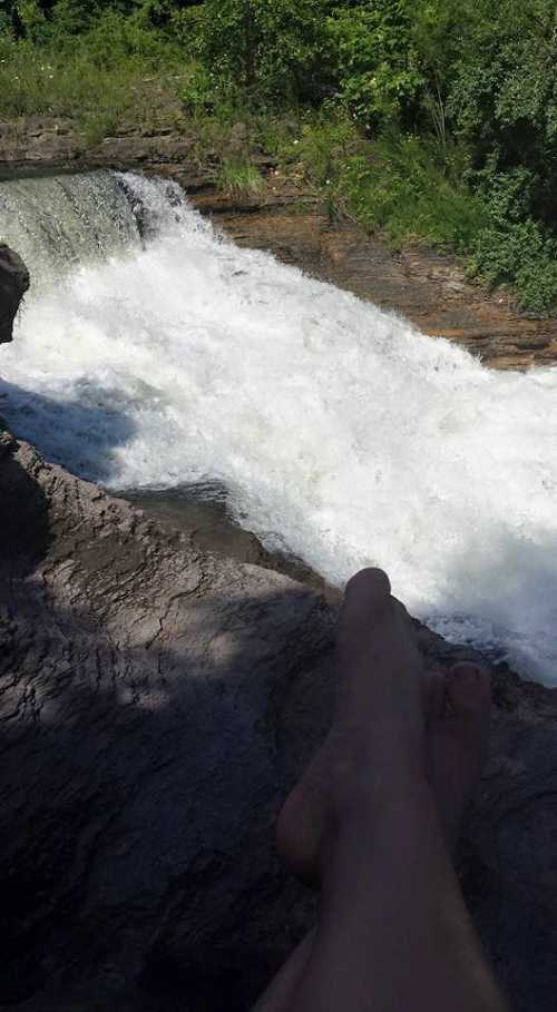 A person's feet resting on a rocky ledge by a rushing waterfall surrounded by lush greenery.