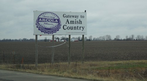 Sign reading "Gateway to Amish Country" with a purple logo, set against a cloudy sky and farmland.