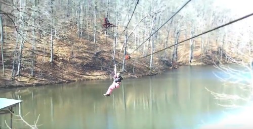 A person zip-lining over a calm lake, surrounded by bare trees in a wooded area.