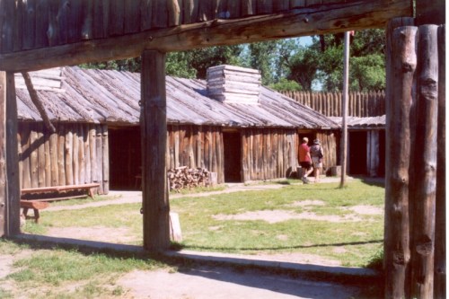A wooden structure with a thatched roof, surrounded by a wooden fence, and two people walking in a grassy area.
