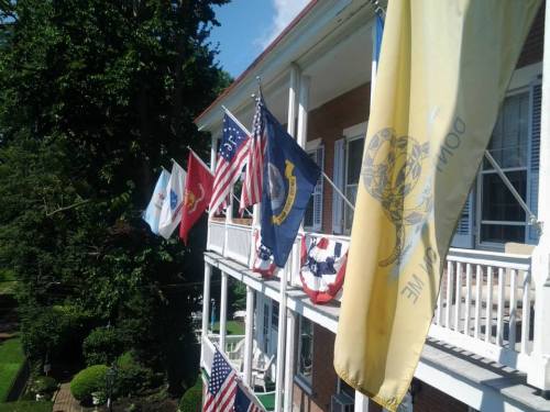 A porch with multiple flags, including U.S. flags and military insignias, surrounded by greenery and blue sky.