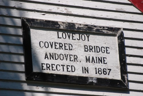 Sign on a building reading "Lovejoy Covered Bridge, Andover, Maine, Erected in 1867" against a white wooden background.