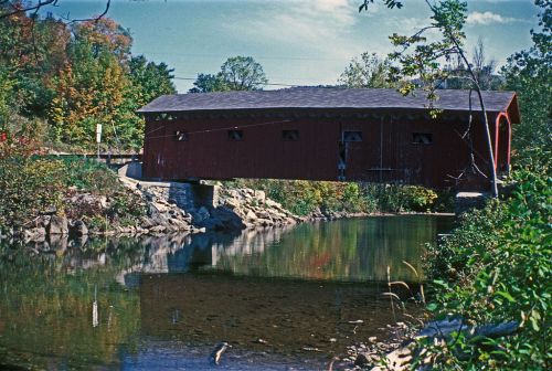 A red covered bridge spans a calm river, surrounded by trees with autumn foliage reflecting in the water.
