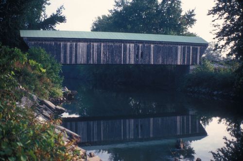 A wooden covered bridge spans a calm river, surrounded by trees and reflecting in the water below.