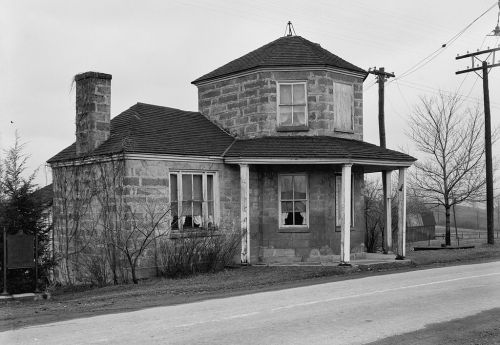 A historic stone house with a unique turret, set along a rural road, surrounded by bare trees and power lines.