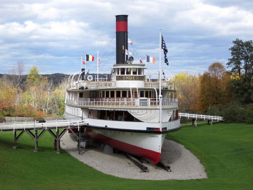 A historic steamboat, the Ticonderoga, sits on land surrounded by trees and flags, with a cloudy sky above.