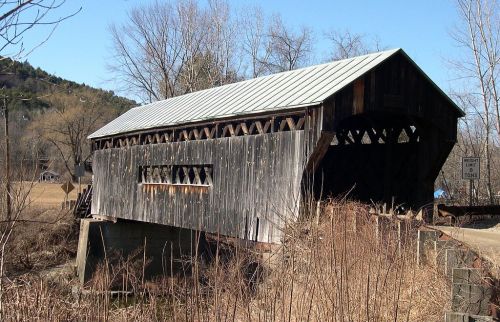 A rustic covered bridge with a wooden structure, surrounded by trees and tall grass under a clear blue sky.
