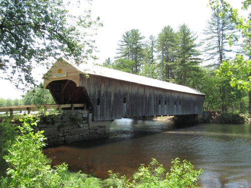 A wooden covered bridge spans a river, surrounded by lush greenery and tall trees under a bright sky.