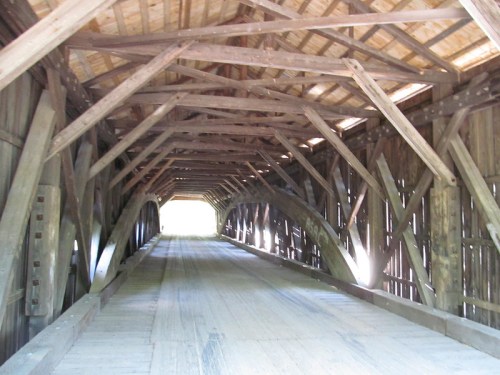 Interior view of a wooden covered bridge, showcasing exposed beams and a light-filled opening at the far end.