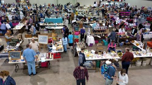 A bustling indoor market with numerous tables filled with various goods and many people browsing and shopping.