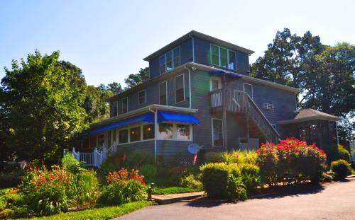 A multi-story blue house with a porch, surrounded by lush greenery and colorful flowers on a sunny day.