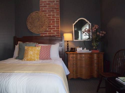 Cozy bedroom with a bed, decorative pillows, a wooden dresser, a mirror, and a lamp, featuring a brick accent wall.