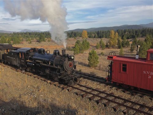 A vintage steam locomotive puffs smoke as it travels along a scenic railway surrounded by trees and mountains.