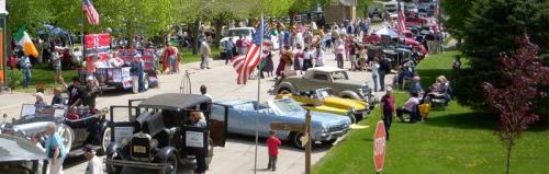 A vibrant car show with vintage vehicles, flags, and crowds enjoying the event in a sunny park setting.