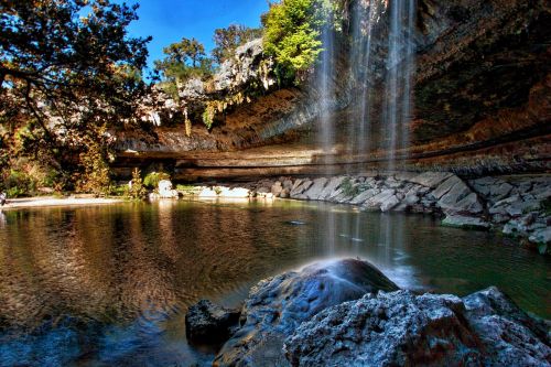 A serene waterfall cascades into a clear pool surrounded by rocky cliffs and lush greenery under a bright blue sky.