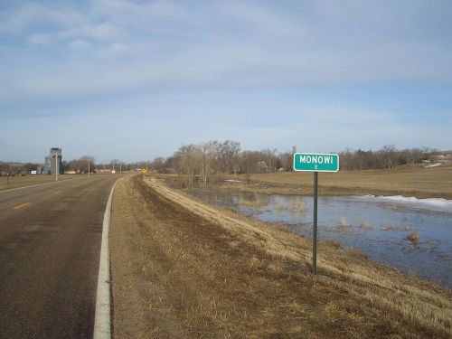 A roadside sign for Monowi, with a rural landscape and a small body of water nearby.