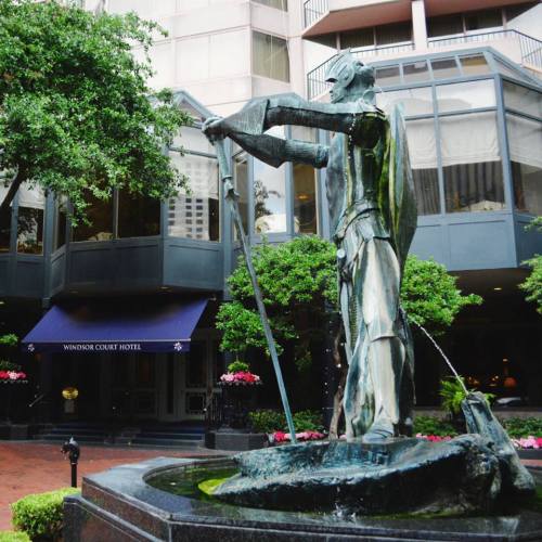 A statue of a man holding a staff stands in front of the Windsor Court Hotel, surrounded by greenery and flowers.