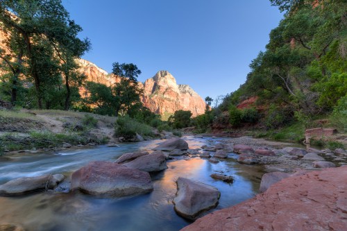 A serene river flows through a canyon, surrounded by lush greenery and towering red rock formations under a clear blue sky.