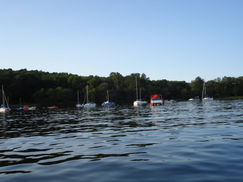A calm lake scene with several boats anchored near a wooded shoreline under a clear blue sky.