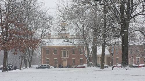A historic brick building surrounded by snow-covered trees in a winter landscape. Snowflakes gently fall.