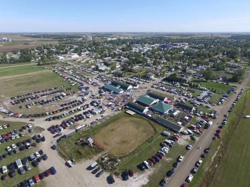 Aerial view of a large outdoor event with crowds and numerous parked cars in a rural town setting.