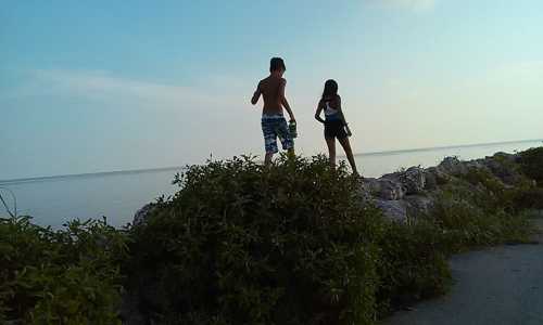 Two children stand on a rocky shore, facing the calm water under a clear sky.