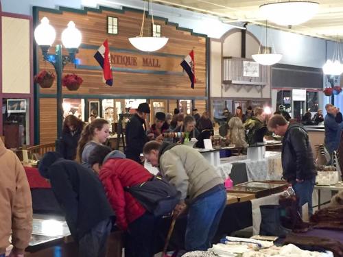 A bustling antique mall with shoppers browsing various items at tables, surrounded by decorative flags and lights.