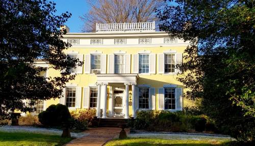 A large yellow historic house with white trim, surrounded by greenery and a clear blue sky.