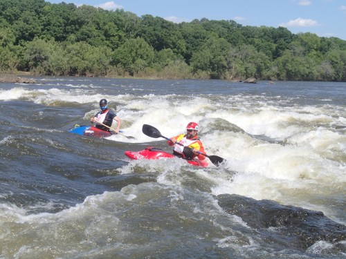 Two kayakers navigate through whitewater rapids on a river, surrounded by lush green trees under a clear blue sky.