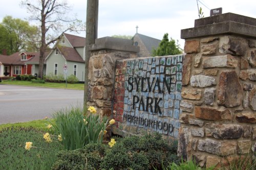A stone sign reading "Sylvan Park Neighborhood" surrounded by flowers and greenery, near a residential area.