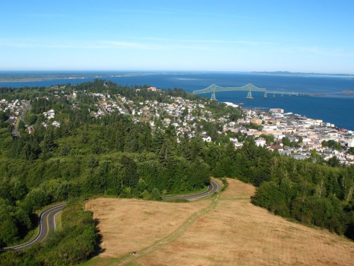 Aerial view of a coastal town with green hills, a winding road, and a bridge over a blue waterway under a clear sky.