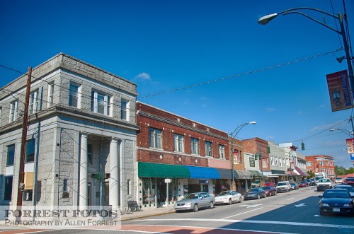 A charming downtown street with historic buildings, shops, and clear blue skies. Cars are parked along the road.