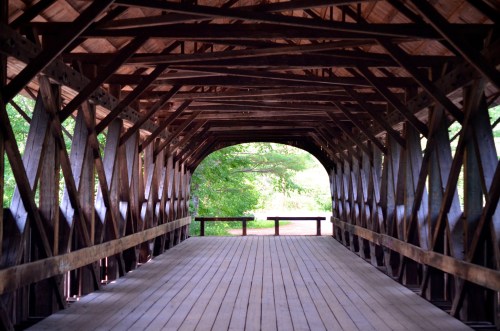 Interior view of a covered wooden bridge, featuring a wooden floor and intricate wooden supports, with greenery visible outside.