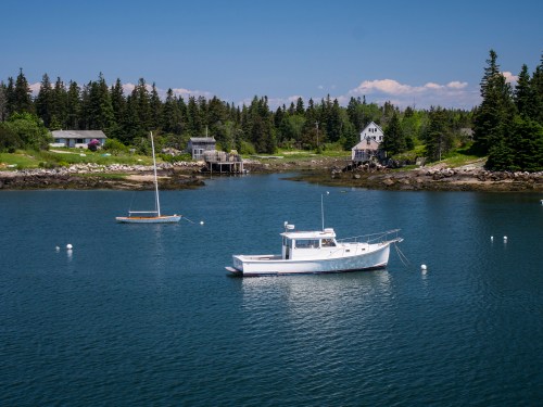 A serene coastal scene with two boats anchored in calm water, surrounded by lush trees and charming houses.