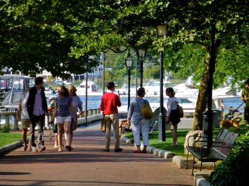 People stroll along a tree-lined walkway by the water, with boats visible in the background and benches nearby.