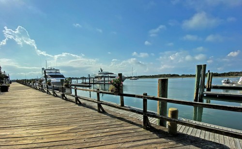 A serene marina scene with boats docked, wooden walkways, and a clear blue sky with scattered clouds.