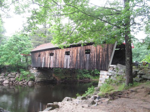 A wooden covered bridge spans a calm river, surrounded by lush green trees and rocky banks.