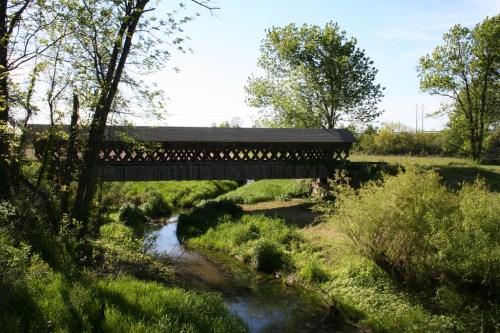 A covered wooden bridge spans a small stream, surrounded by lush greenery and trees under a clear blue sky.