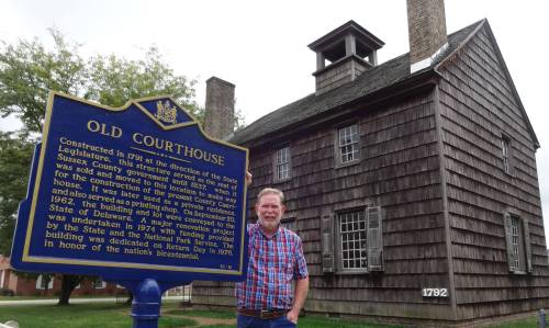 A man stands beside a historical marker in front of a wooden courthouse building with a year "1792" displayed.