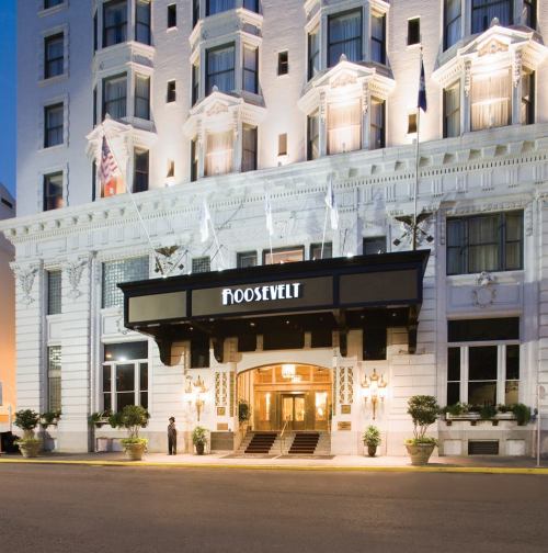 The Roosevelt Hotel entrance, featuring elegant architecture and flags, with a doorman standing by the entrance.