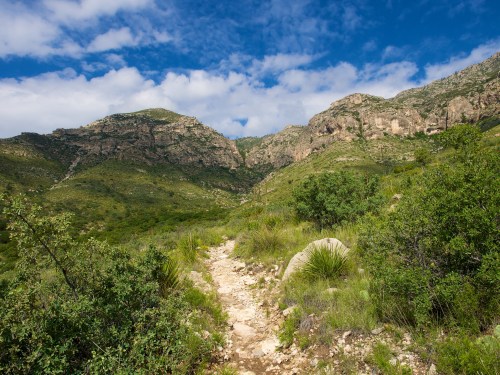 A winding dirt path leads through lush greenery and rocky hills under a partly cloudy sky.