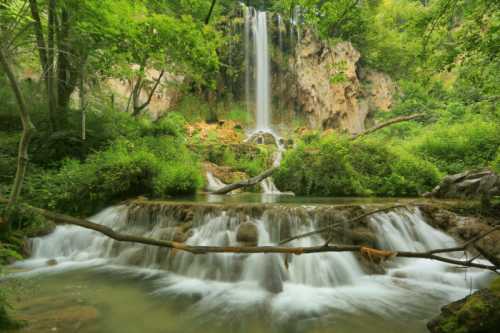 A serene waterfall cascades into a lush green landscape, surrounded by trees and rocky formations.